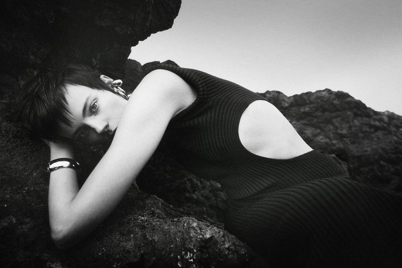 Monochrome fashion shot of a woman in a dark dress reclining against rocks with a sky background, showcasing minimalist jewelry.