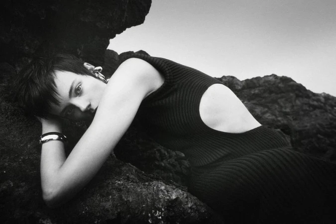 Monochrome fashion shot of a woman in a dark dress reclining against rocks with a sky background, showcasing minimalist jewelry.
