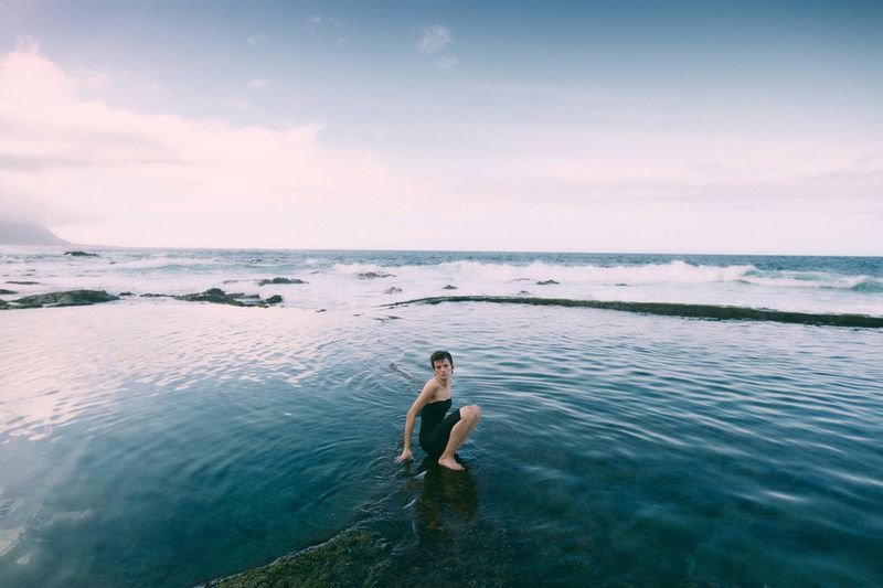 Woman squats on rocks in a shallow ocean pool on a clear day. Ocean waves stretch to the horizon under a blue and white sky.