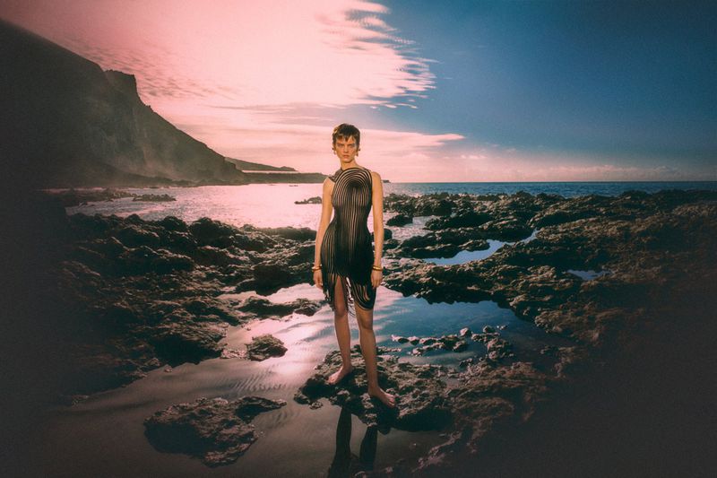 A stylish adult female in a unique dress poses on a rocky beach with ocean and cliffs under a blue and pink sky.