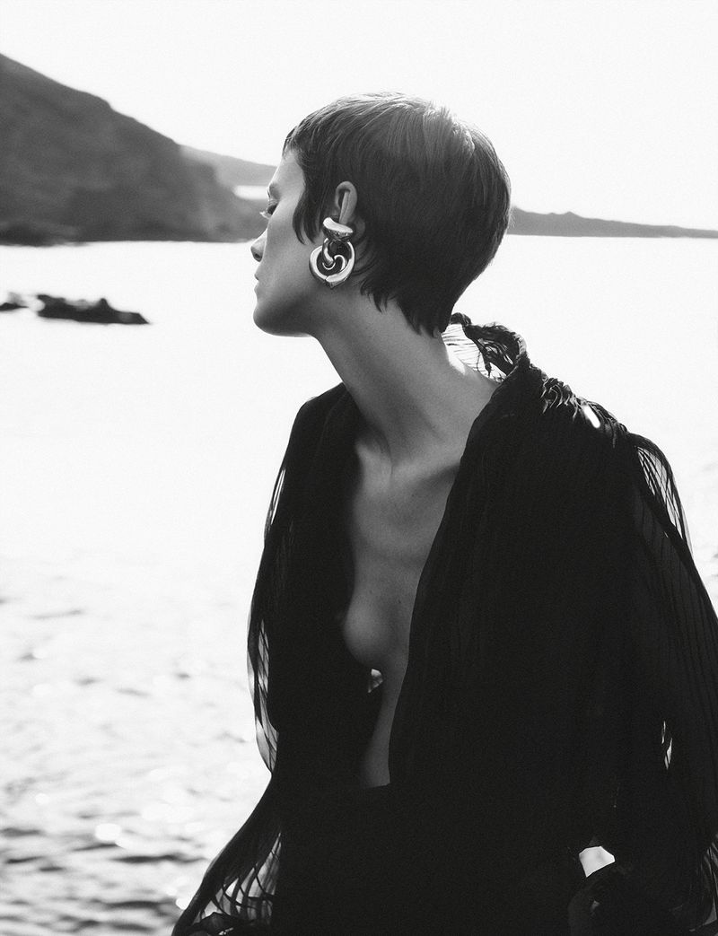 Black and white portrait of woman with short hair wearing large earrings and black robe, outdoors on coastline with mountain backdrop.