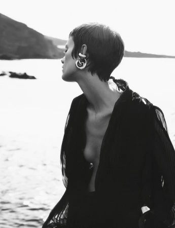 Black and white portrait of woman with short hair wearing large earrings and black robe, outdoors on coastline with mountain backdrop.