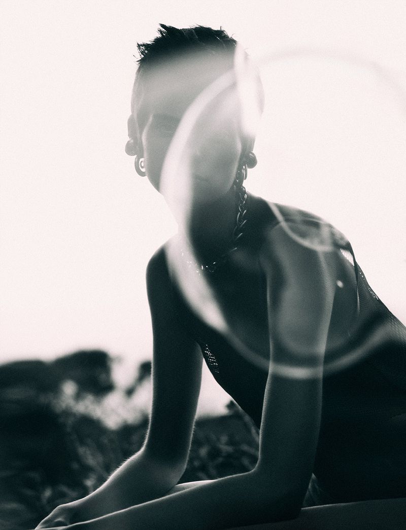 Monochrome portrait of a person with short hair, wearing large earrings and a chain necklace, posing outdoors with sunlight and shadows.