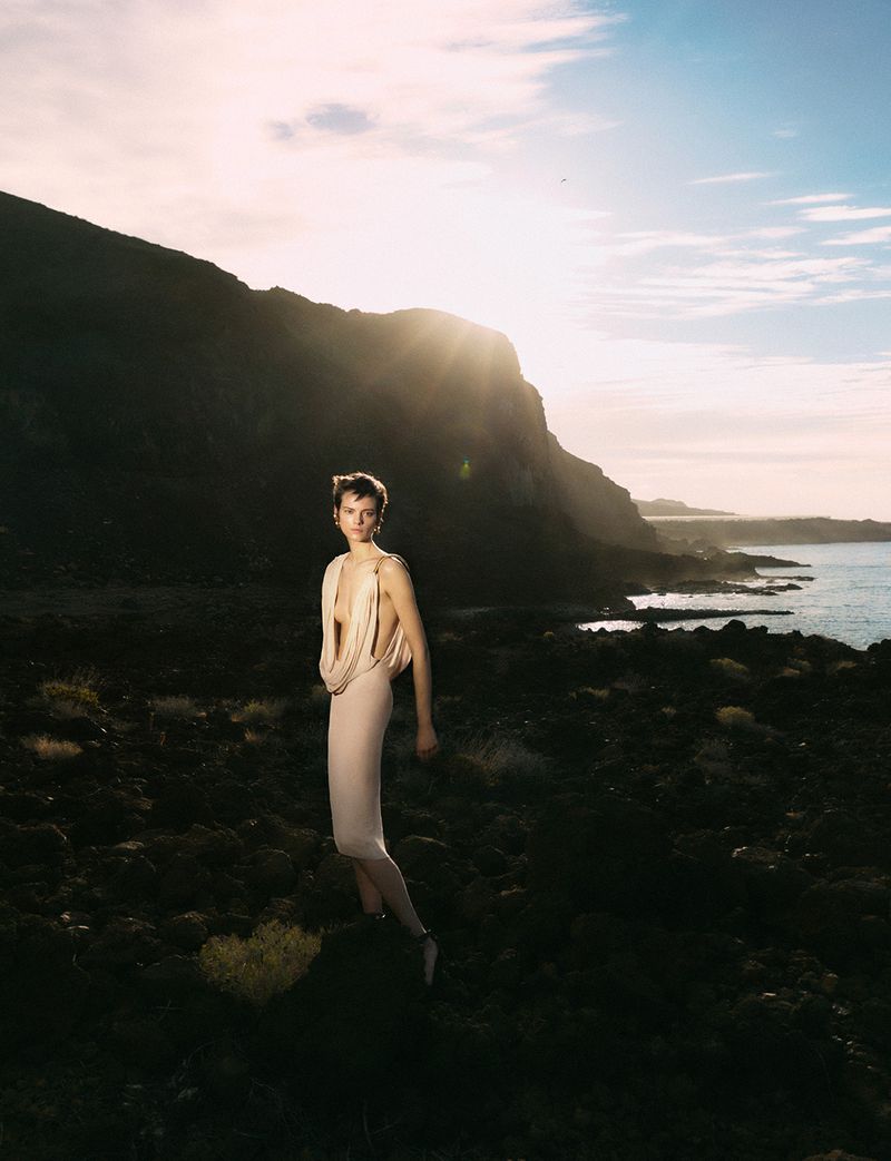 A woman in a draped dress poses in a rocky landscape near the ocean at sunset. The sun glows behind her and a dark mountain.