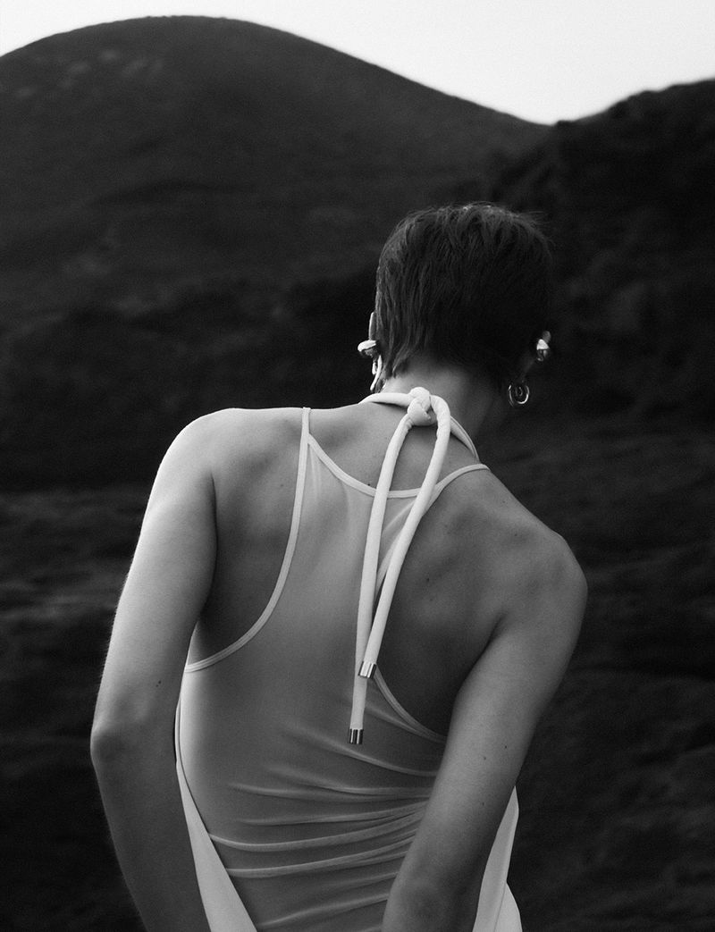 Monochrome portrait from behind of woman with short hair wearing a white swimsuit with mountain landscape in the background, earring.