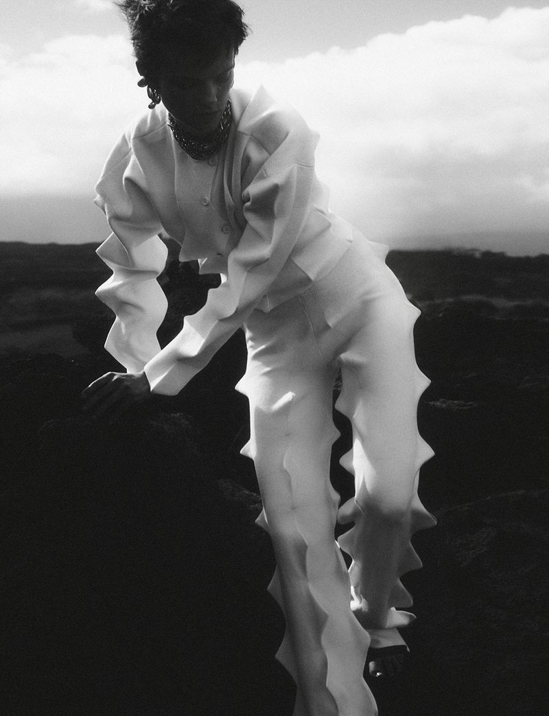 Monochrome fashion shot: adult posing in white textured outfit on dark terrain under cloudy sky. Haute couture editorial style.