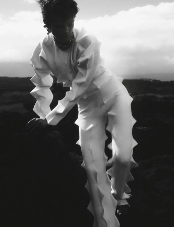 Monochrome fashion shot: adult posing in white textured outfit on dark terrain under cloudy sky. Haute couture editorial style.