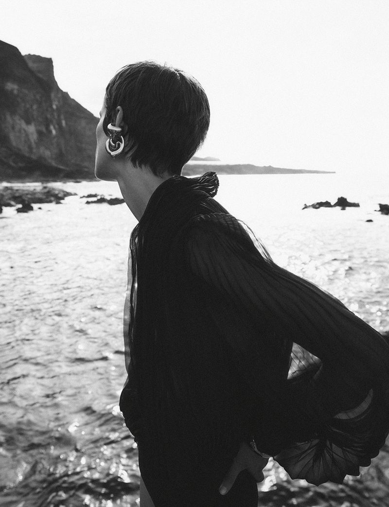 Monochrome outdoor portrait of a young adult woman with short hair and hoop earrings looking out over a rocky shoreline and ocean.