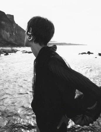 Monochrome outdoor portrait of a young adult woman with short hair and hoop earrings looking out over a rocky shoreline and ocean.
