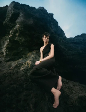 Maaike Klaasen sitting barefoot on dark rocky crags in a black one-shoulder knit dress with gold jewellery for Numéro Switzerland.