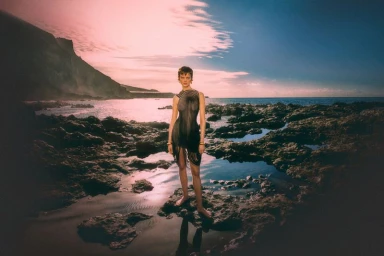 Model standing on wet dark rocks beside the ocean under a glowing sunset sky for Numero Switzerland