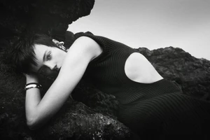 Model with dark hair wearing a black sleeveless dress reclining against dark rocky coast formations in a monochrome fashion image