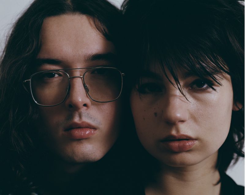 Close-up studio portrait of a young adult man with long hair and eyeglasses and a young adult woman with short dark hair.