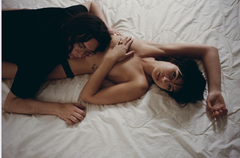 A shirtless woman lying on a white bedsheet is embraced by a man with glasses and dark hair, creating a relaxed and intimate scene.