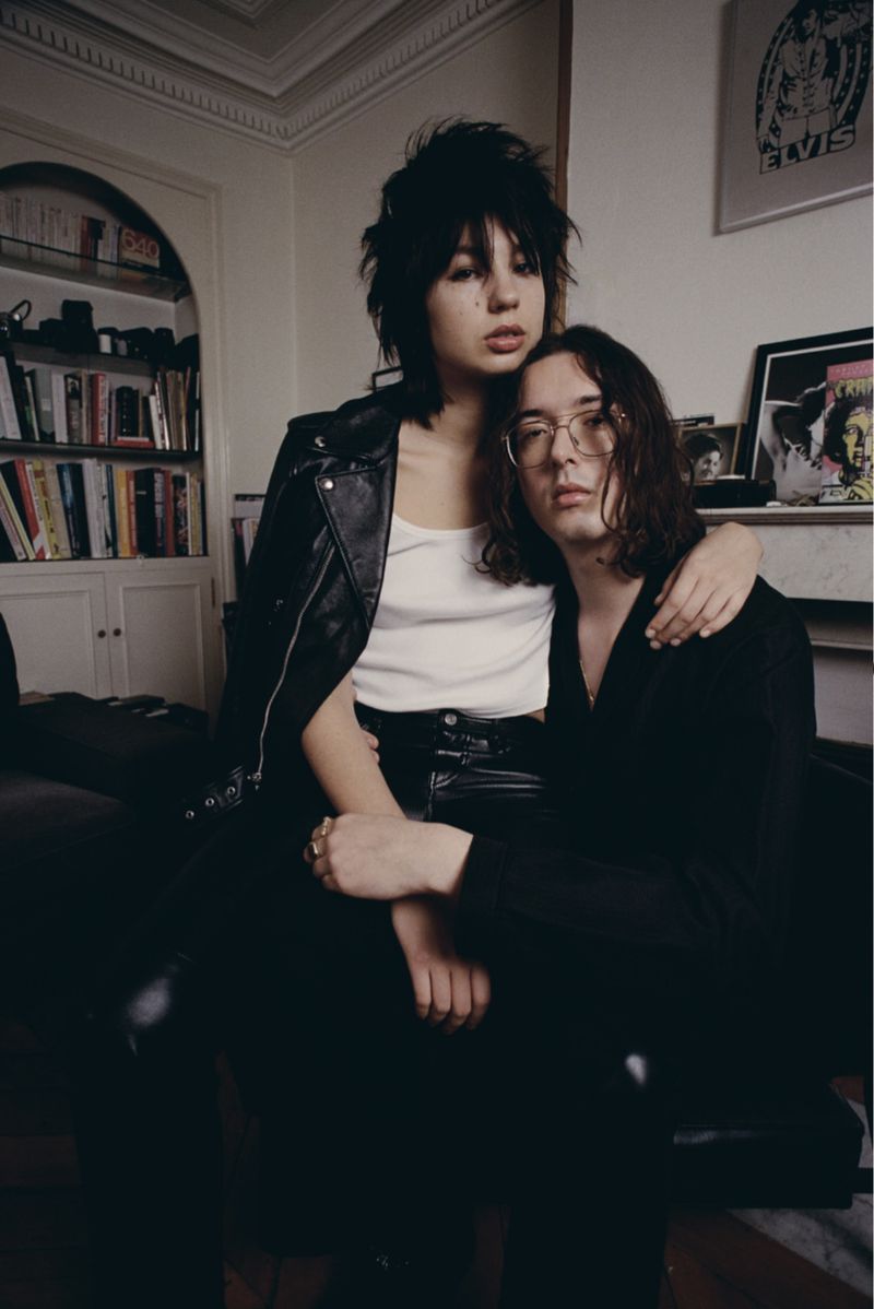 Indoor portrait of a stylish couple: a young woman with spiky dark hair and a young man with long hair, posing in casual black clothing.