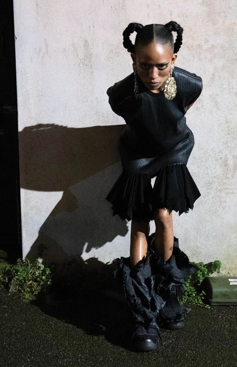 Young person in a black dress and deconstructed boots poses against a textured wall, showcasing avant-garde fashion with a spotlight effect.