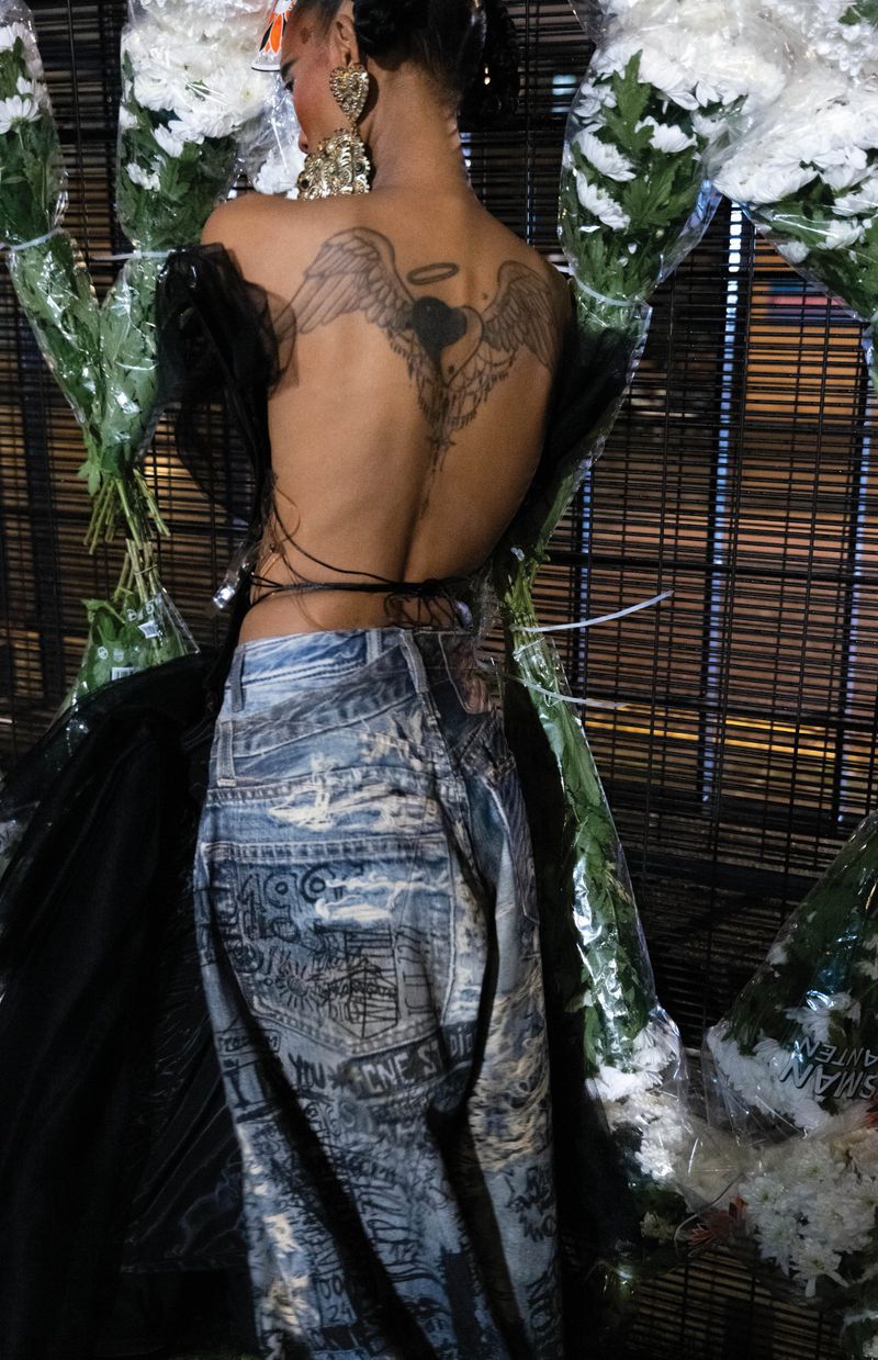 Adult woman poses with a tattoo on her back, wearing jeans and standing near white flower bouquets in an indoor environment.