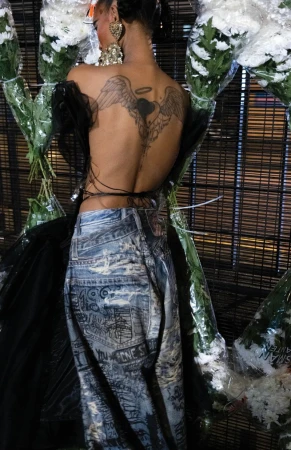 Adult woman poses with a tattoo on her back, wearing jeans and standing near white flower bouquets in an indoor environment.