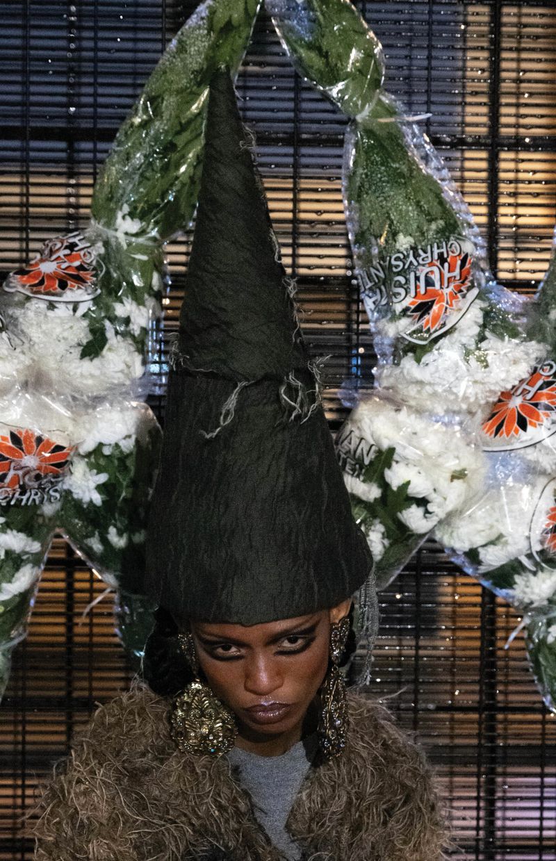 Fashion portrait: person wearing cone-shaped hat and large earrings posing in front of patterned background with floral arrangements.