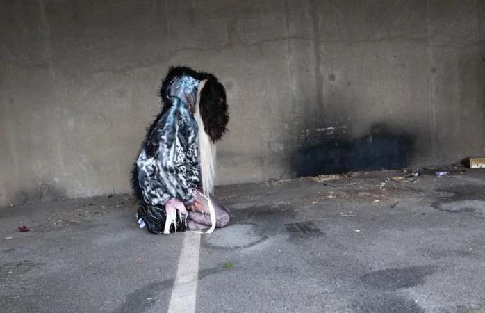 Model Adesuwa Aighewi kneeling on asphalt in an oversized printed hooded jacket against a raw concrete wall.