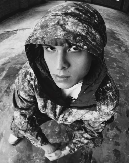 Monochromatic close-up portrait of a young adult wearing a hooded camouflage jacket indoors, looking upwards at the camera.