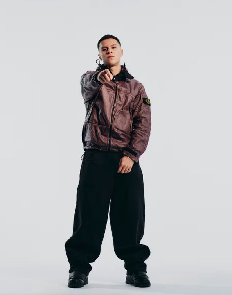 Young adult male in a brown jacket and black pants points at the viewer in a studio setting with a white background and neutral colors.
