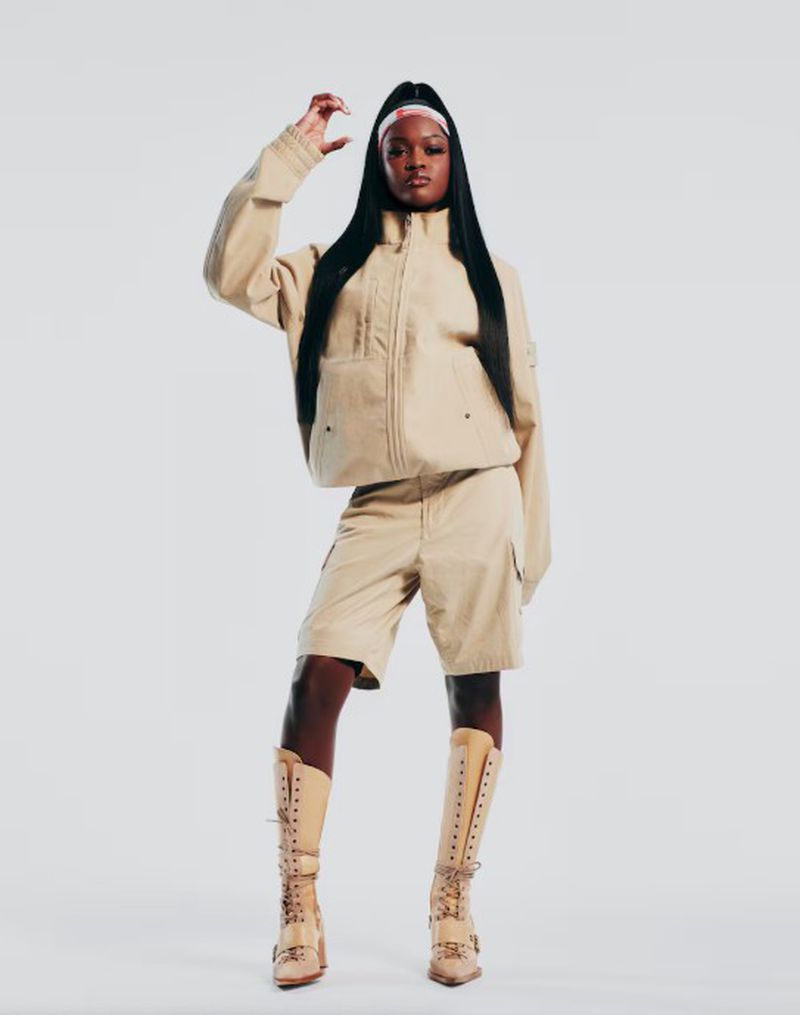 A full shot of an adult woman posing in a beige jacket, shorts, and boots, smiling confidently in front of a white backdrop.