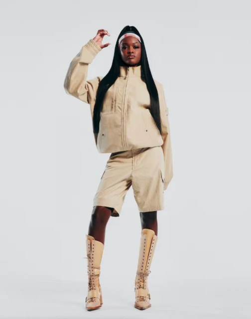 A full shot of an adult woman posing in a beige jacket, shorts, and boots, smiling confidently in front of a white backdrop.