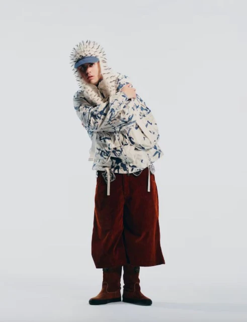 Person in spiked hood and brown culottes strikes a pose in a fashion forward photo studio shot with a white background and medium framing.
