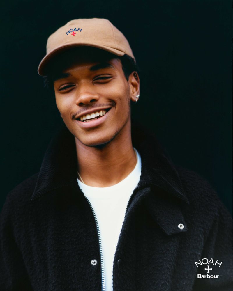 Portrait of a young Black man smiling, wearing a beige cap and black jacket, standing against a dark background. Brand logos visible.