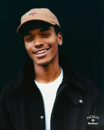 Portrait of a young Black man smiling, wearing a beige cap and black jacket, standing against a dark background. Brand logos visible.