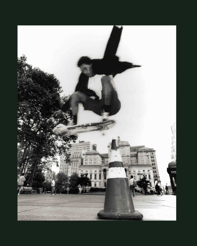 Monochromatic action shot of skateboarder performing aerial trick over traffic cone in urban square with vintage buildings.