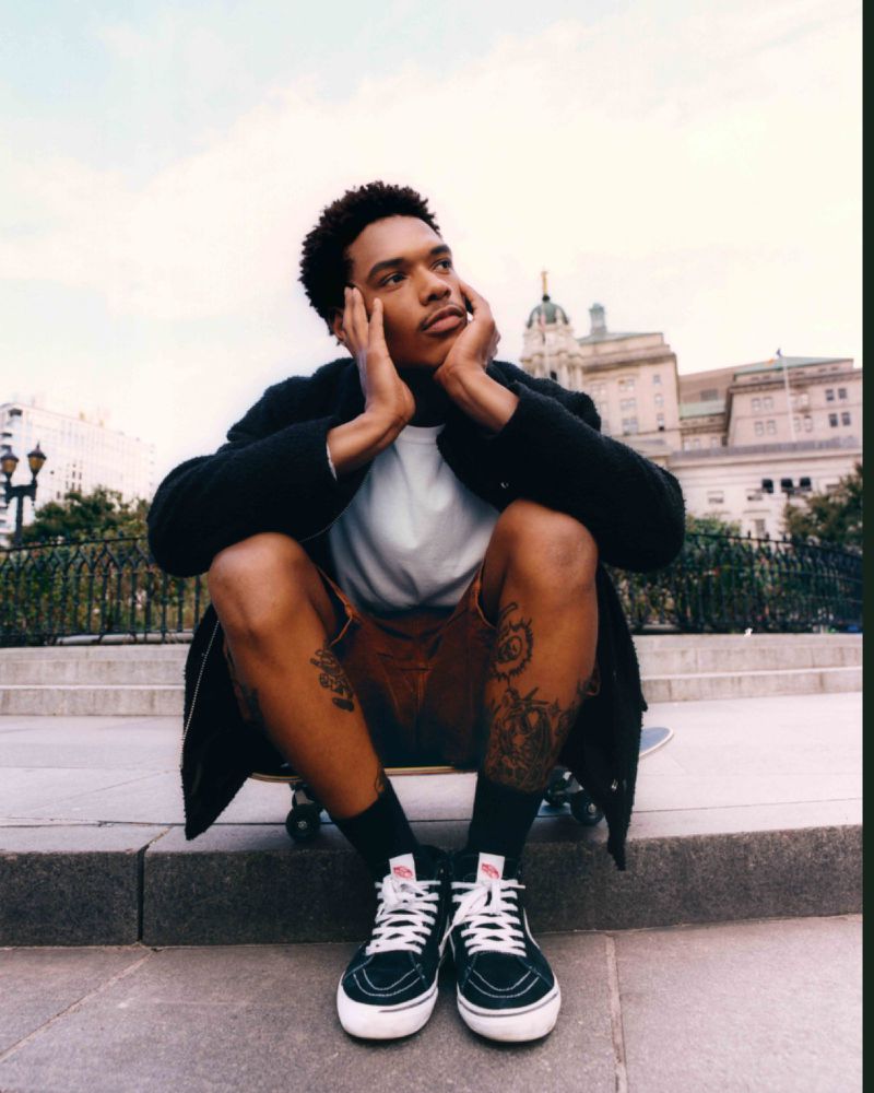 Young man sits on skateboard in city square with European-style buildings in background, wearing shorts, black jacket, and Vans sneakers.