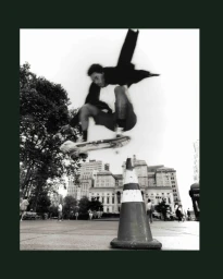 Monochrome action shot of a skateboarder mid-air jumping over a traffic cone in a public square with classical buildings in the background.