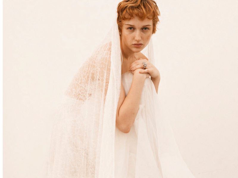 Portrait of a young adult woman with short red hair, wearing a white veil and looking at the camera against a neutral background.