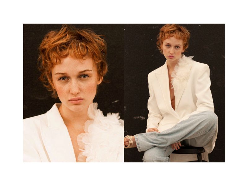Portrait of a young woman with red hair wearing a white blazer and light blue jeans against a dark background in a fashion editorial style.