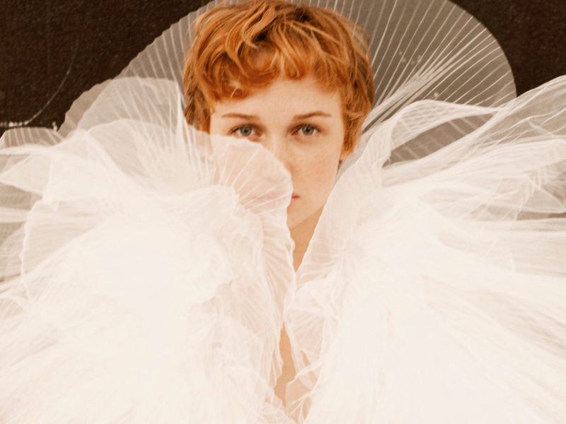 Close-up portrait of a woman with short red hair, wearing a white tulle collar, set against a dark, textured backdrop, fashion photography.