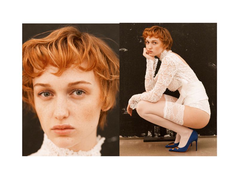 Portrait of a woman with short auburn hair wearing a white lace top and high heels, posing against a dark background in studio lighting.