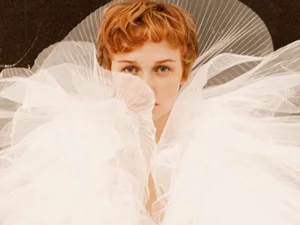 Close-up portrait of a woman with short reddish hair surrounded by an exaggerated white pleated tulle ruff against a dark backdrop.