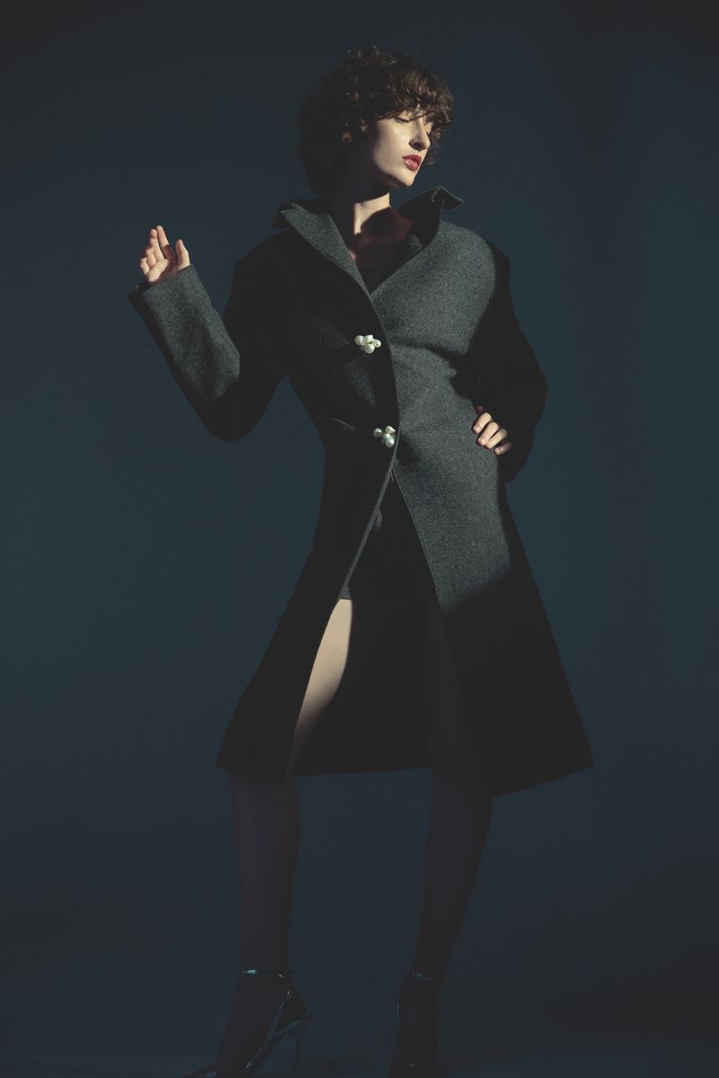A woman poses in a long, gray coat with pearl buttons, standing in a dimly lit studio against a dark blue backdrop, evoking high fashion.