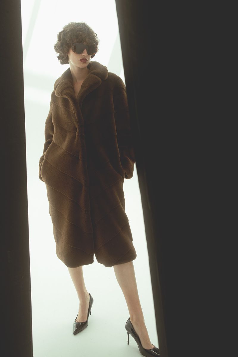 A model poses in a brown faux fur coat with black heels, dark glasses, and a neutral background in a fashion photography shot.