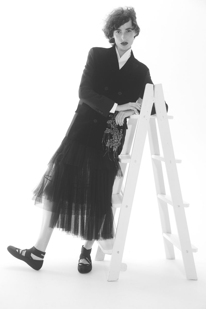 Black and white fashion shot of a person in a jacket and tulle skirt leaning against a white ladder in a bright photo studio.