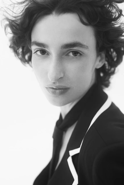 Monochrome portrait of a woman with curly hair, wearing a dark blazer with white trim, against a plain white backdrop.