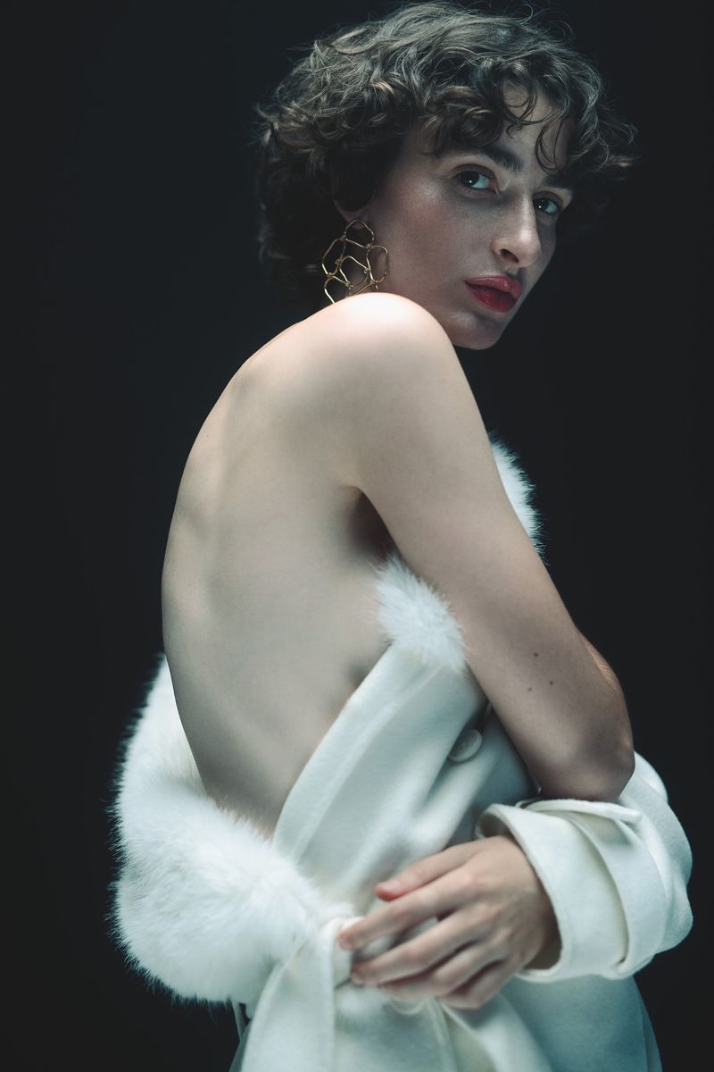 Woman with short dark hair poses in white coat with fur trim in front of dark background, gazing over her shoulder.