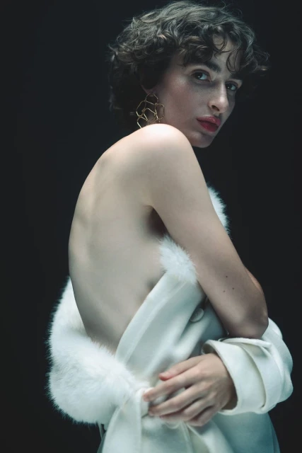Woman with short dark hair poses in white coat with fur trim in front of dark background, gazing over her shoulder.