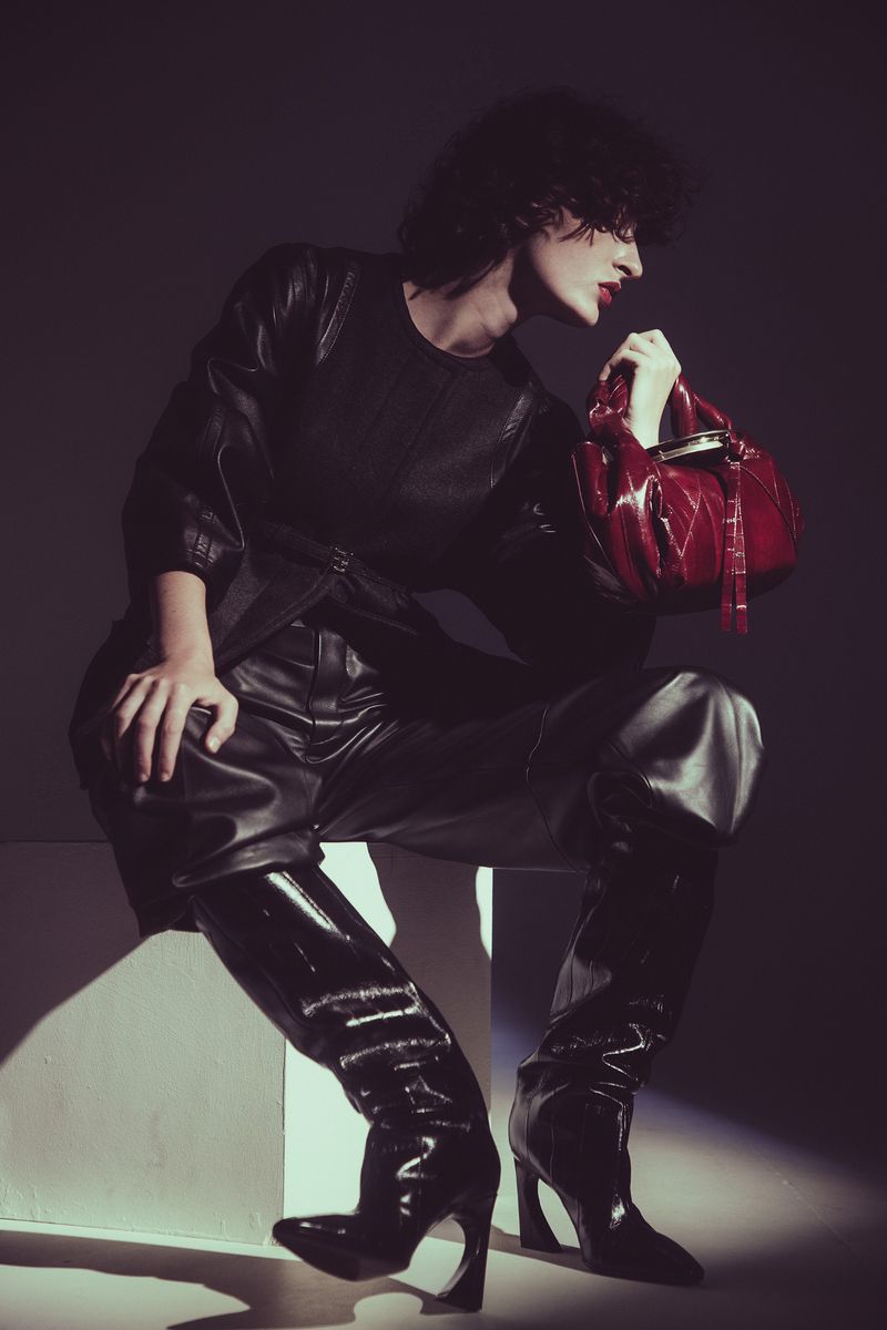 A person in a black leather outfit poses with a red handbag against a dark background, seated on a white block in a studio setting.