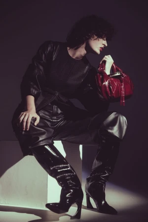 A person in a black leather outfit poses with a red handbag against a dark background, seated on a white block in a studio setting.
