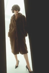 Model wearing dark sunglasses and a long brown coat framed by dark vertical foreground shapes against a bright white background.
