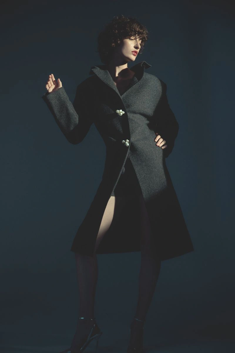 Model in a structured dark grey coat with ornamental buttons posing against a deep moody backdrop.