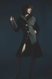 Model in a structured dark grey coat with ornamental buttons posing against a deep moody backdrop.
